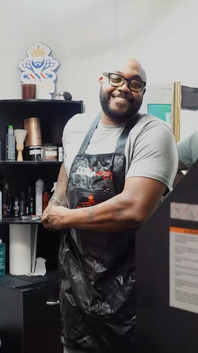 The founder smiling while tying on his barber apron beside a shelf of grooming products